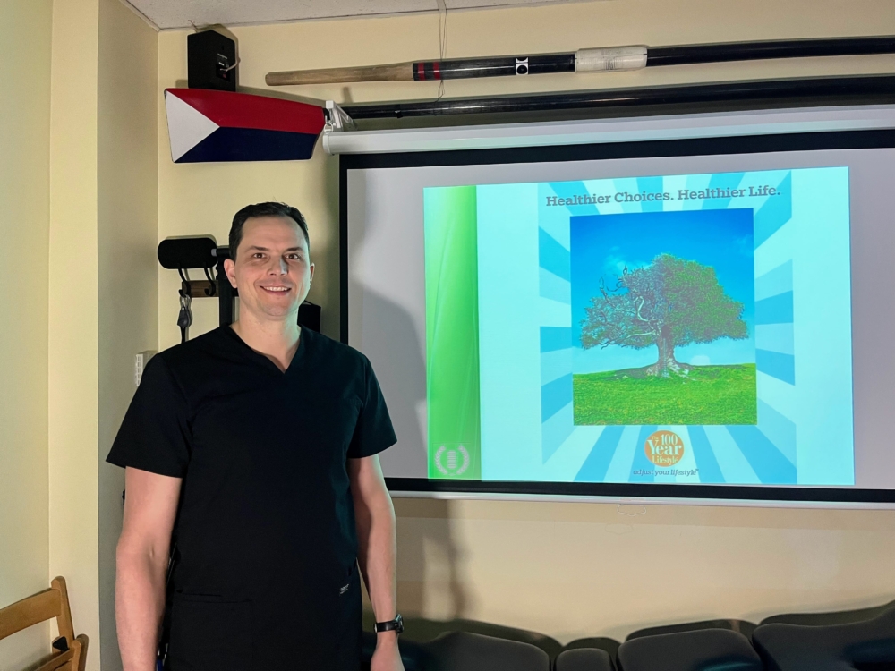 A man in black scrubs stands smiling next to a projector screen displaying an image of a tree with the text “Healthier Choices. Healthier Life.” Rowing equipment is mounted on the wall above.