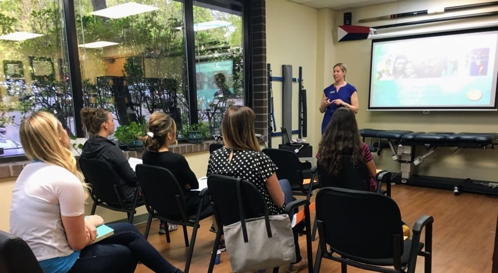 Anna Cummins stands at the front of the clinic giving a presentation on chiropractic to five seated women. A projector screen displays slides, and large windows let in natural light. Notebooks and bags are on chairs and laps.