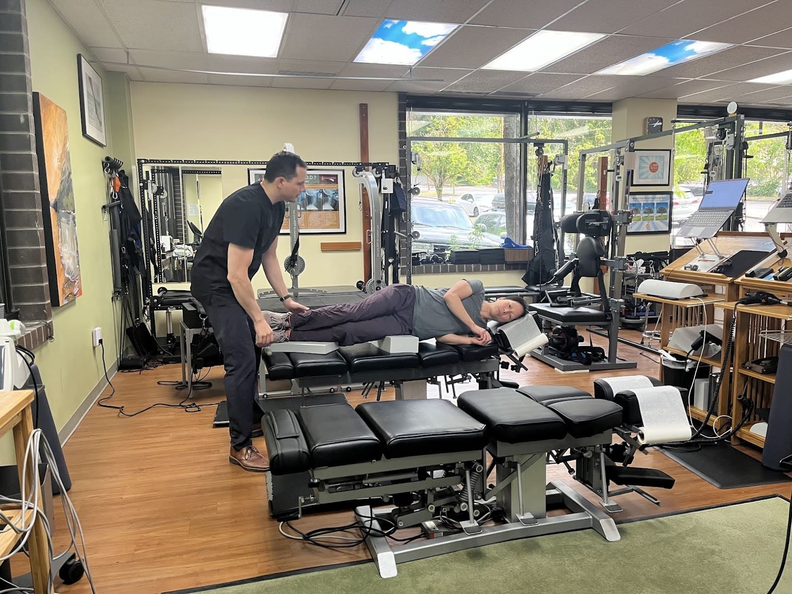 A chiropractor stands beside a patient lying on their side on an adjustable treatment table in a well-equipped clinic, preparing for a hyperkyphosis treatment. Exercise machines and large windows are visible in the background.