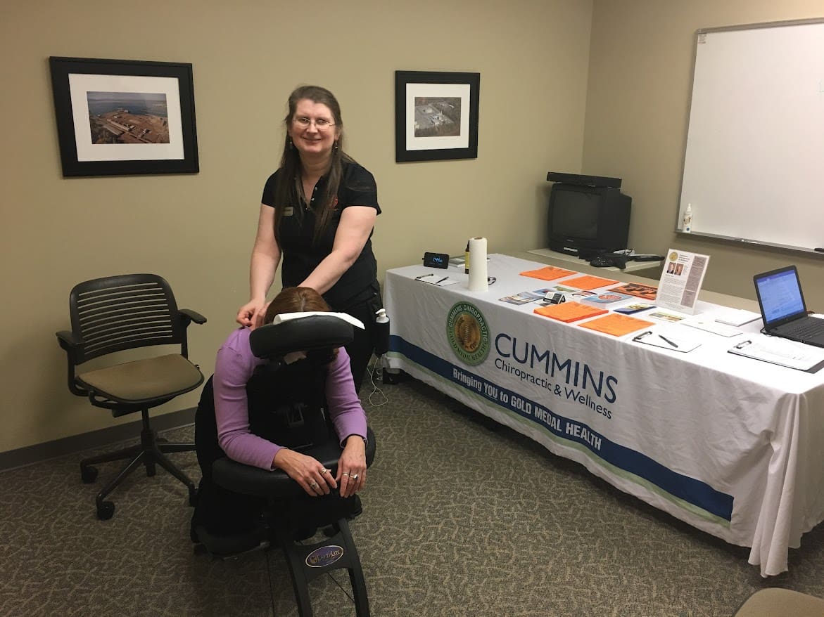 A woman gives a seated massage to another woman in an office, highlighting Bellevue massage therapy. Behind them, brochures, a laptop, and a banner for Cummins Chiropractic & Wellness are displayed. Framed photos hang on the wall.