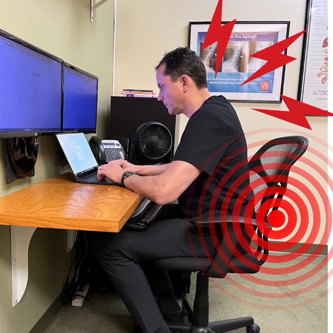 A man sits at a desk working on a laptop with two monitors on the wall. Red stress lines and bullseye graphics highlight his lower back, suggesting discomfort that may benefit from Postural Distortion Treatment.