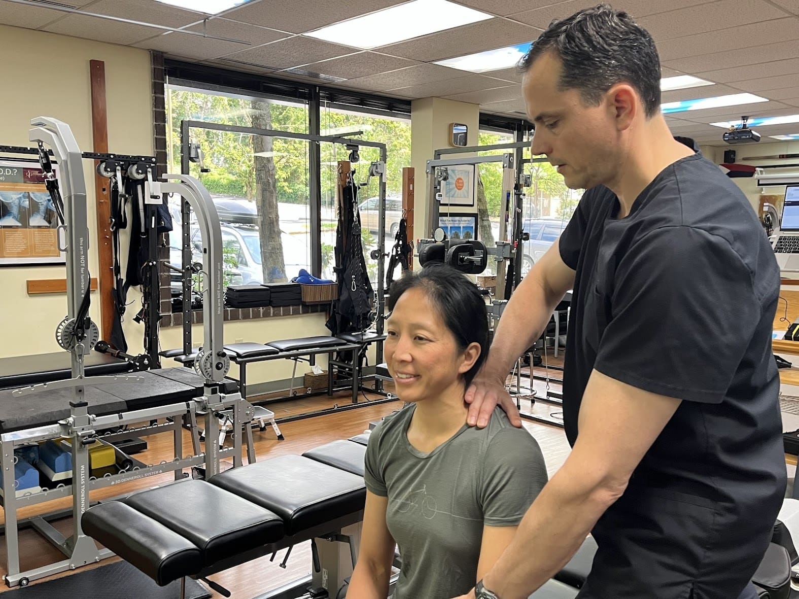 A person sits on a treatment table in a patient center, smiling, while a healthcare professional in black scrubs places his hands on her shoulders to assess or treat her. Exercise equipment is visible in the background.