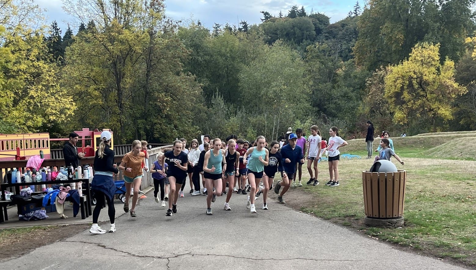 A group of young runners start a race on a paved path in a park, with spectators and a table of water bottles nearby. Trees with green and yellow leaves are in the background.