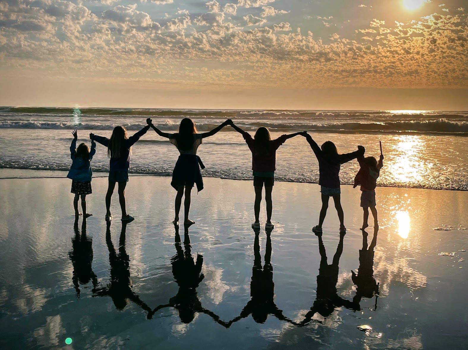 Six people stand hand in hand on a beach at sunset, their silhouettes and reflections visible on wet sand. The serene scene evokes harmony and balance—key elements often sought in Postural Distortion Treatment. Waves and clouds glow behind them.