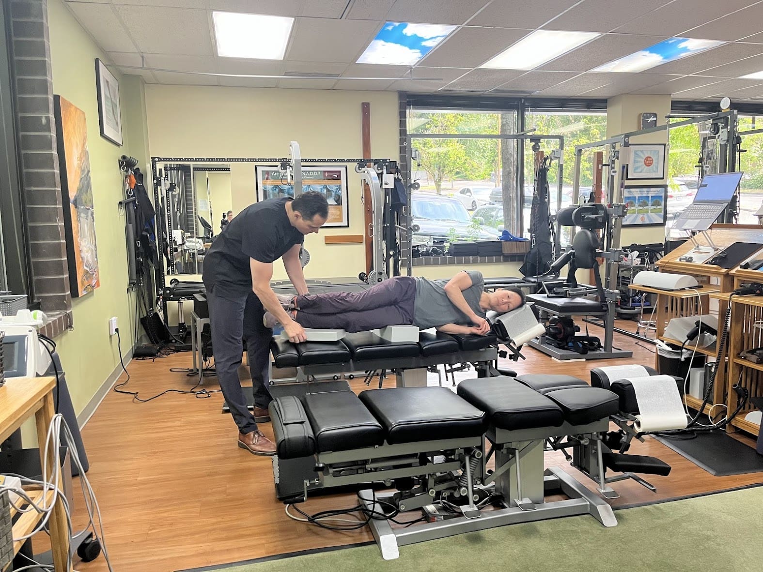 A chiropractor provides chiropractic care to a patient lying on their side on a treatment table in a well-lit clinic with large windows, exercise equipment, and wooden floors.