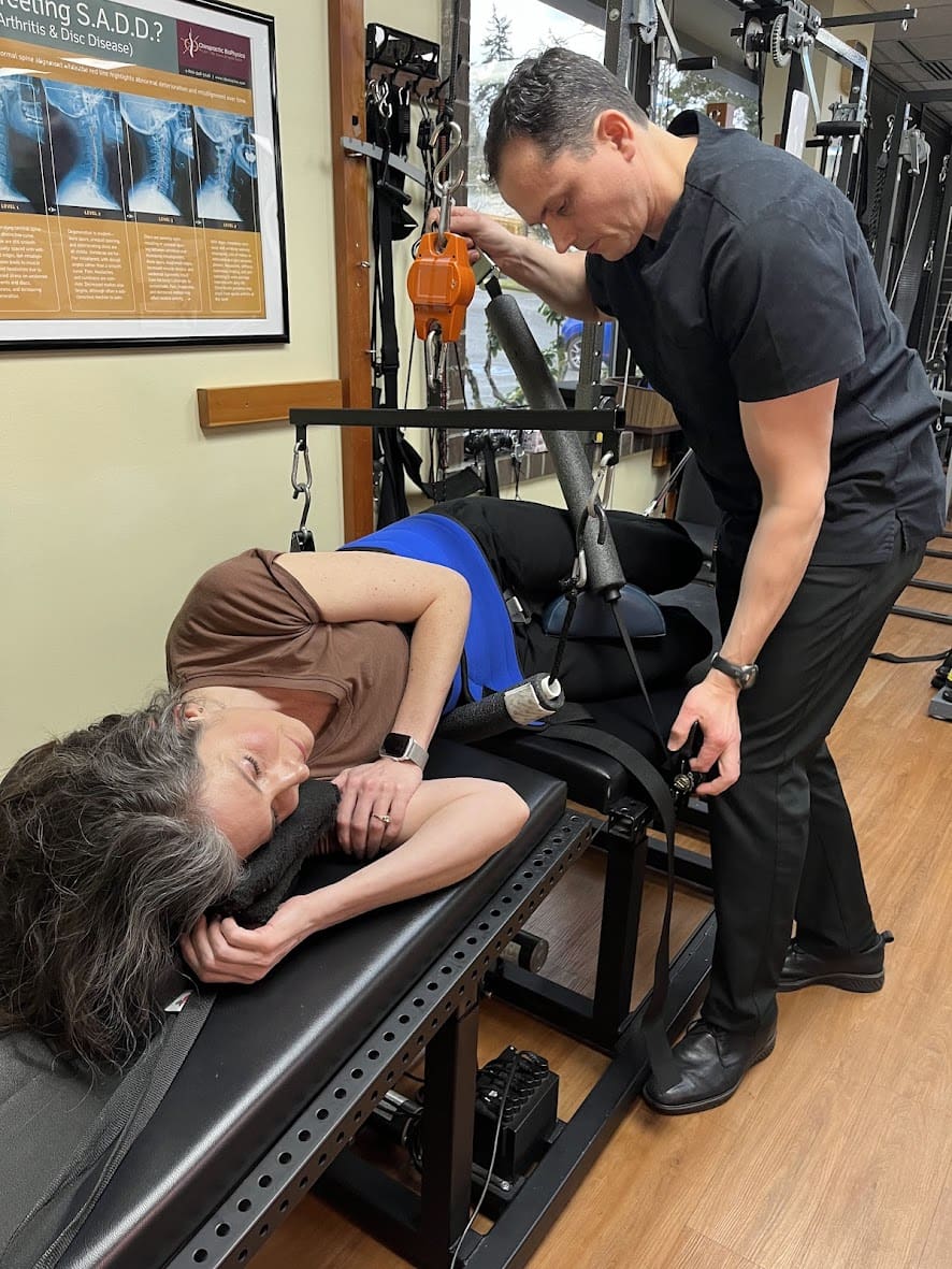 A woman lies on her side on a therapy table for lower back pain treatment, attached to a traction device, while a therapist in black adjusts the equipment in a clinical setting. The woman appears relaxed during the session.