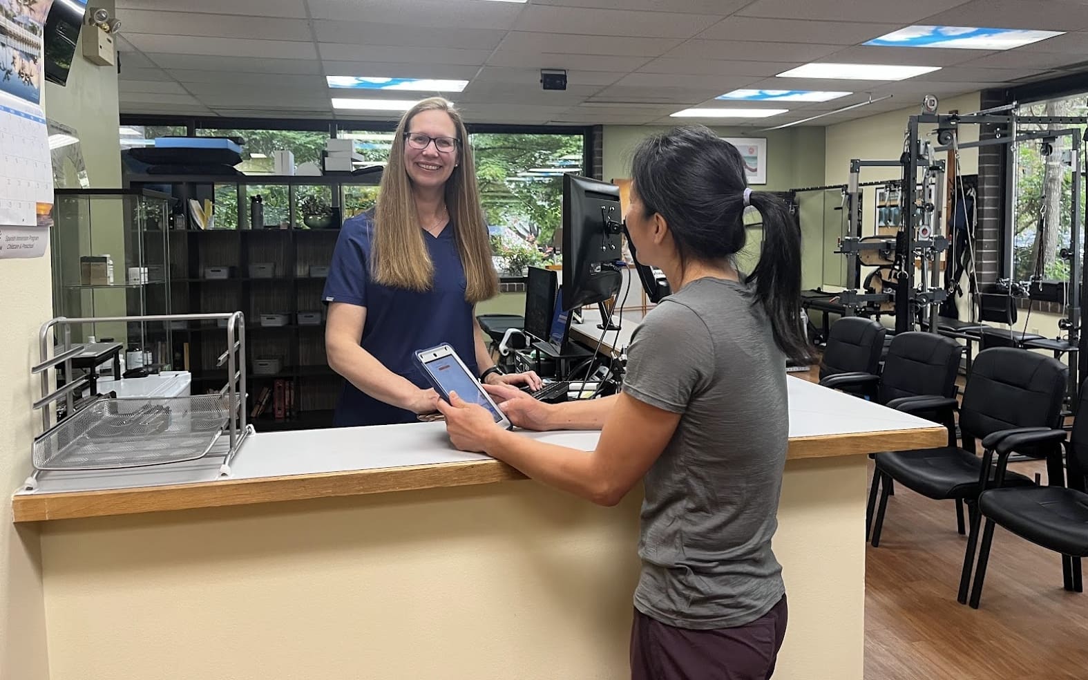 A woman stands at a patient center reception desk holding a phone, speaking with a smiling receptionist in a medical or fitness office. Chairs and exercise equipment are visible in the background.