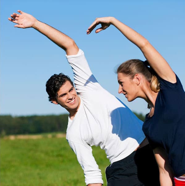 Two people, embracing the 100 year lifestyle, are stretching outdoors on a sunny day. Side by side, they smile and lean sideways with one arm raised above their heads against a vibrant blue sky.