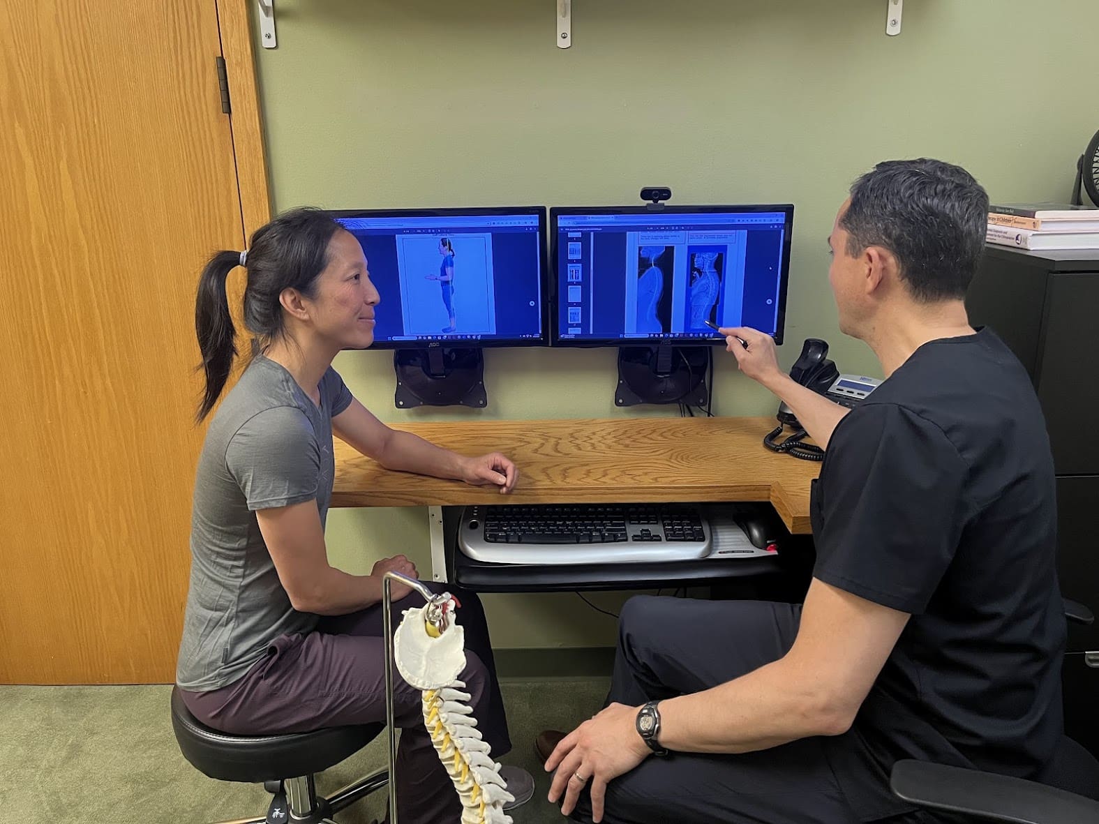 A doctor and a patient sit at a desk reviewing spinal X-ray images on two screens. The doctor points out details related to herniated disc treatment, while a spine model rests on the desk for explanation.