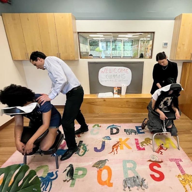 Two people receive chair massages from two massage therapists in an office setting. A colorful alphabet rug covers the floor, while a sign on the window says “Welcome Cummins Corporate Wellness,” reflecting our vision for workplace well-being.