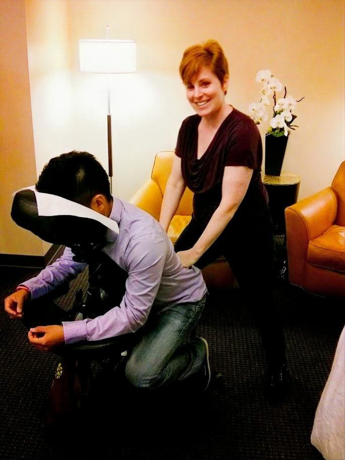A woman smiles while giving a seated massage to a man using a massage chair in a cozy room, reflecting our vision of comfort and relaxation, with an armchair, lamp, and a vase of white flowers in the background.