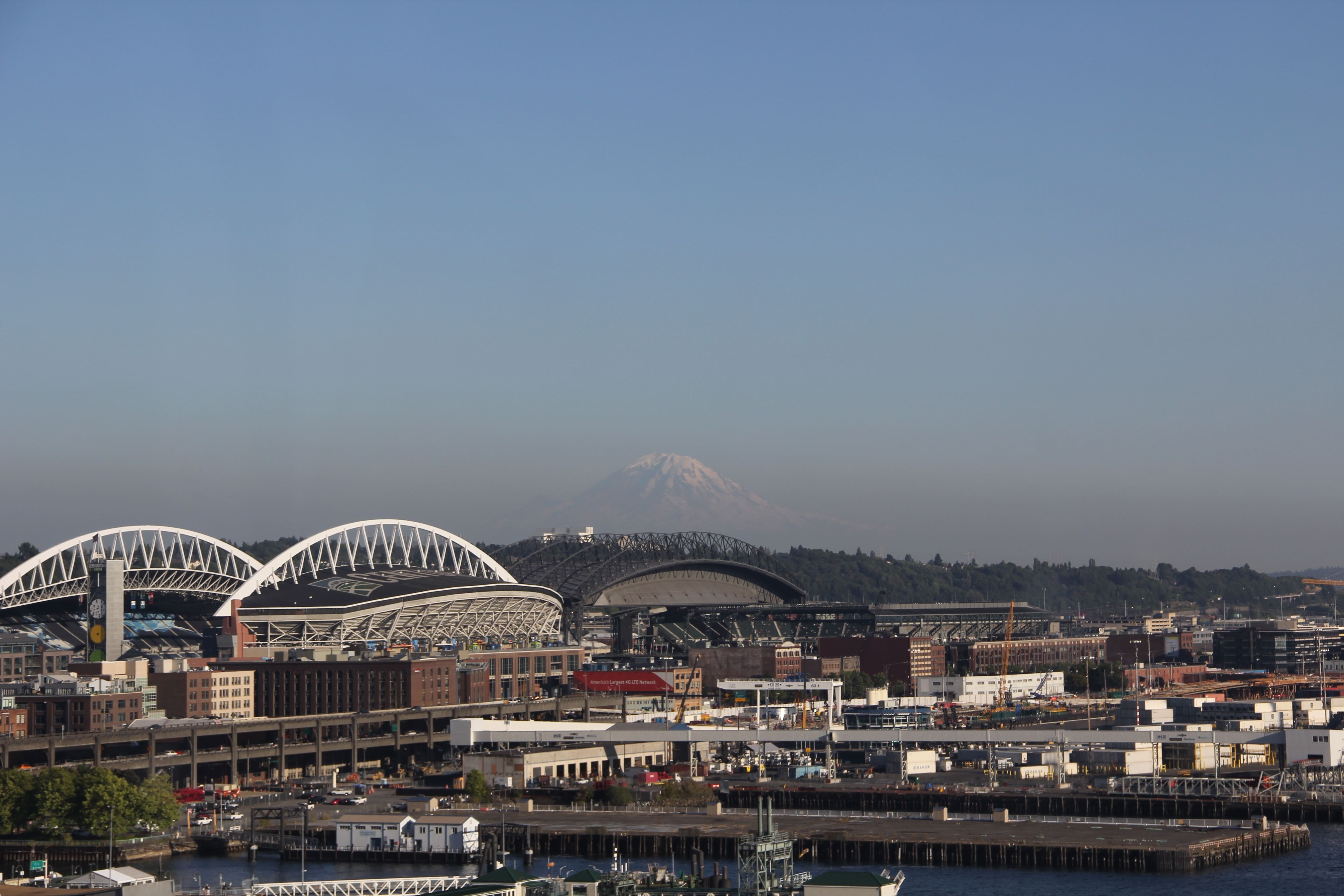 A cityscape featuring two large stadiums and industrial buildings in the foreground, with a snow-capped mountain visible in the hazy distance under a clear blue sky—a setting that inspires both movement and posture improvement.