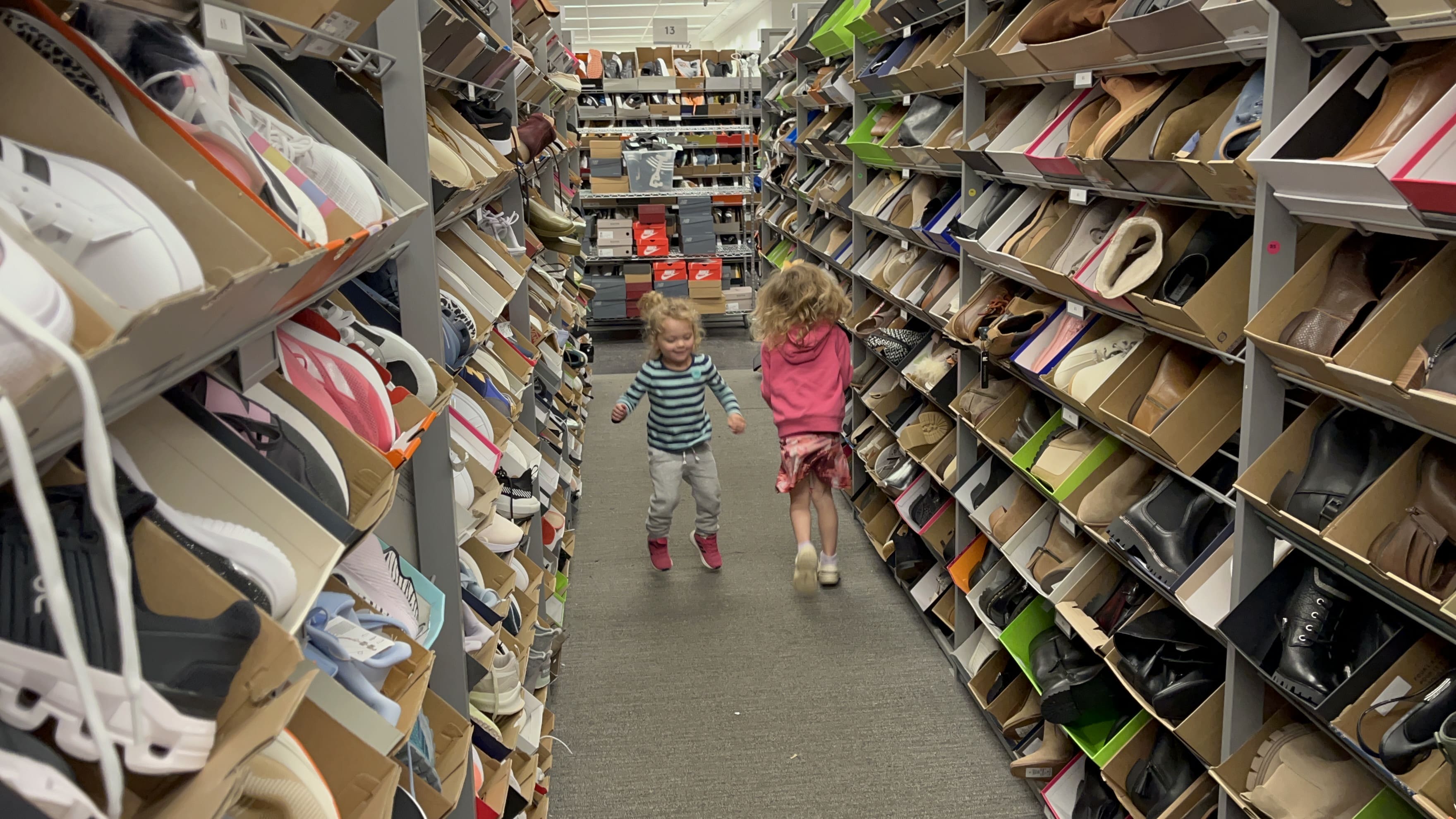 Two young children are running down an aisle in a shoe store near Cummins Factoria Chiropractic office, surrounded by shelves filled with various types of shoes on both sides. The scene is bright and lively.