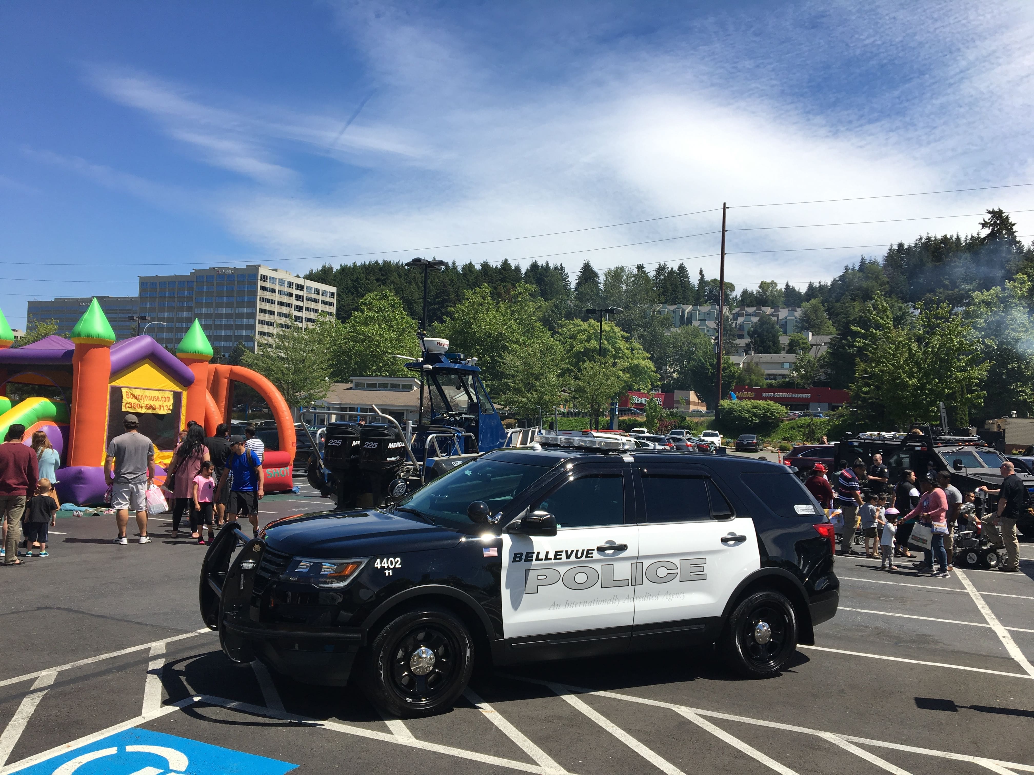 A Bellevue Police SUV is parked in a lot near a crowd, a helicopter, colorful inflatables, at a lively community event on a sunny day. Trees and buildings provide the backdrop.
