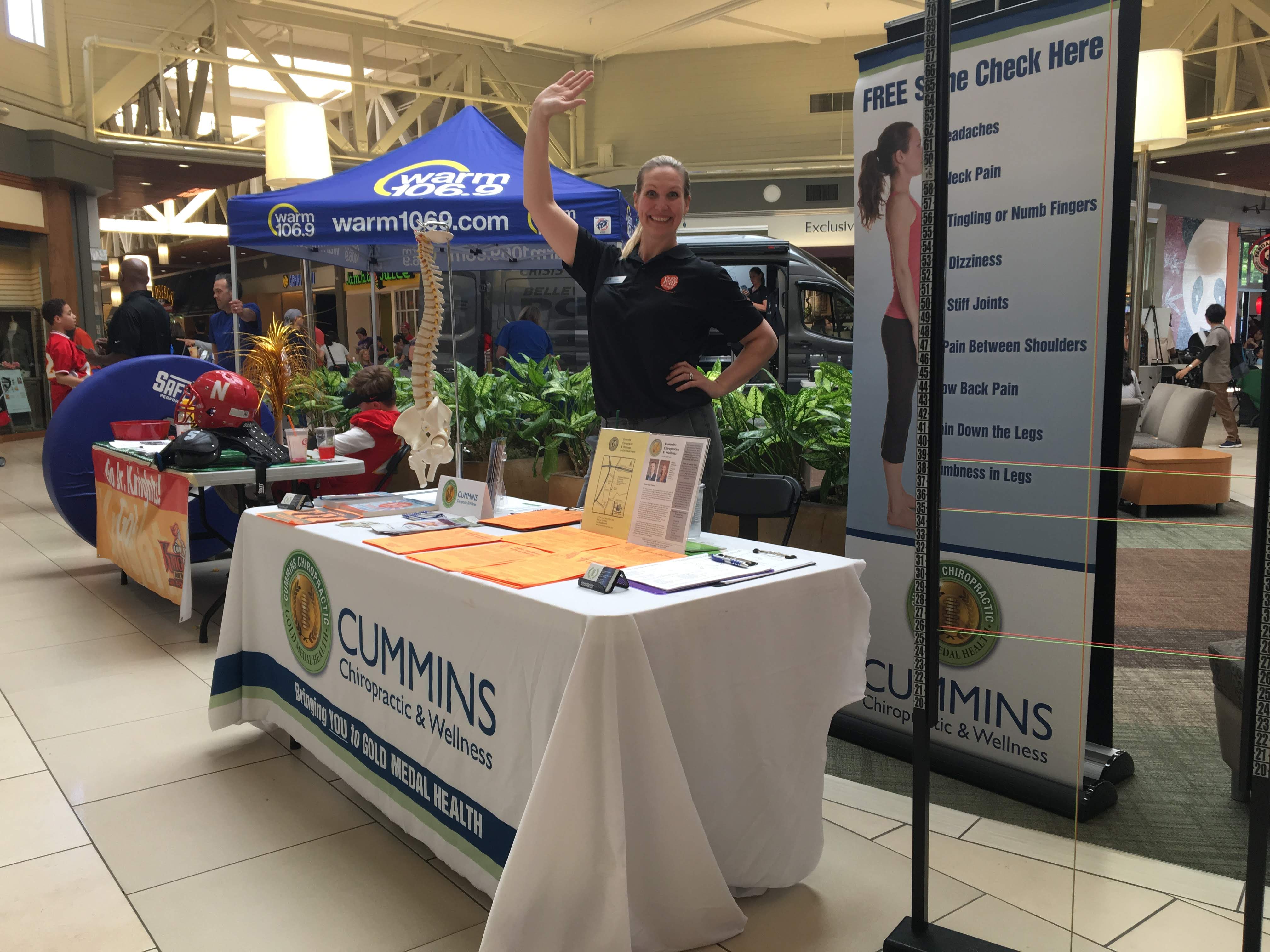 A woman stands smiling and waving behind a Cummins Chiropractic & Wellness table at a mall health fair, with informational pamphlets, a spine model, and a “Free Spine Check Here” sign displayed—showcasing Factoria chiropractic care. Other booths are visible in the background.