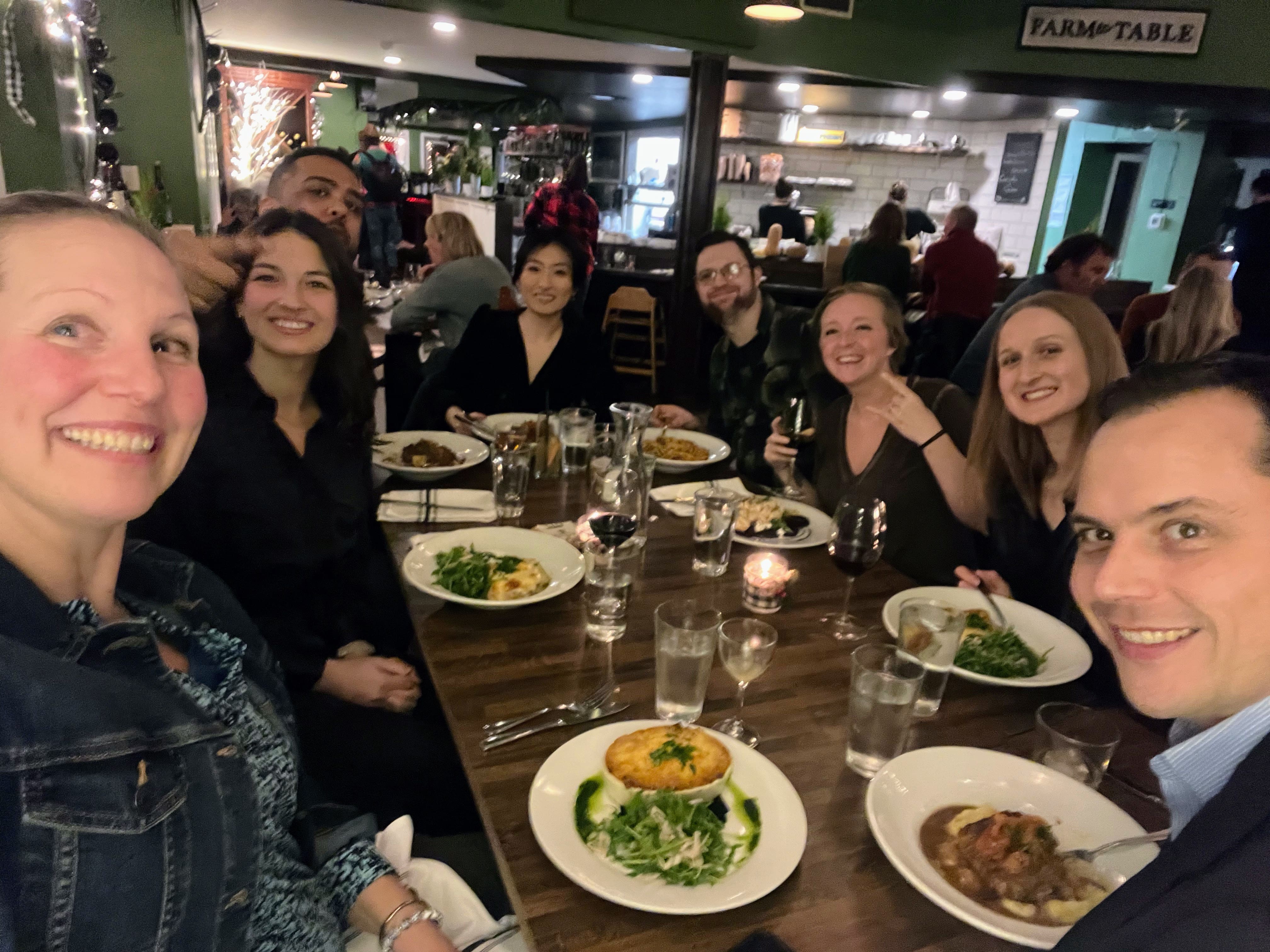 Eight people smiling and posing for a group selfie around a wooden table in an Issaquah restaurant, with plates of food and drinks in front of them; the atmosphere is warm and casual.