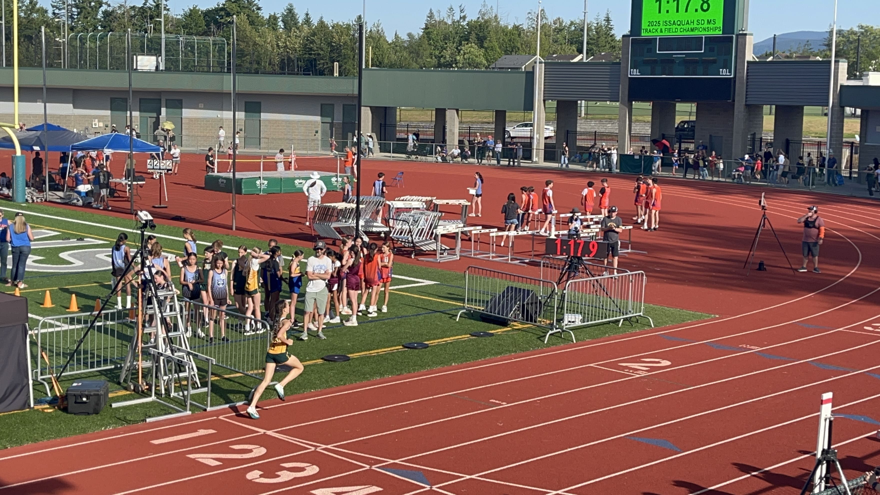 A runner in black and yellow crosses the finish line on the Issaquah outdoor track, while other athletes, officials, and spectators watch. A digital timer above the track reads 1:17.8. It's a sunny day with trees in the background.
