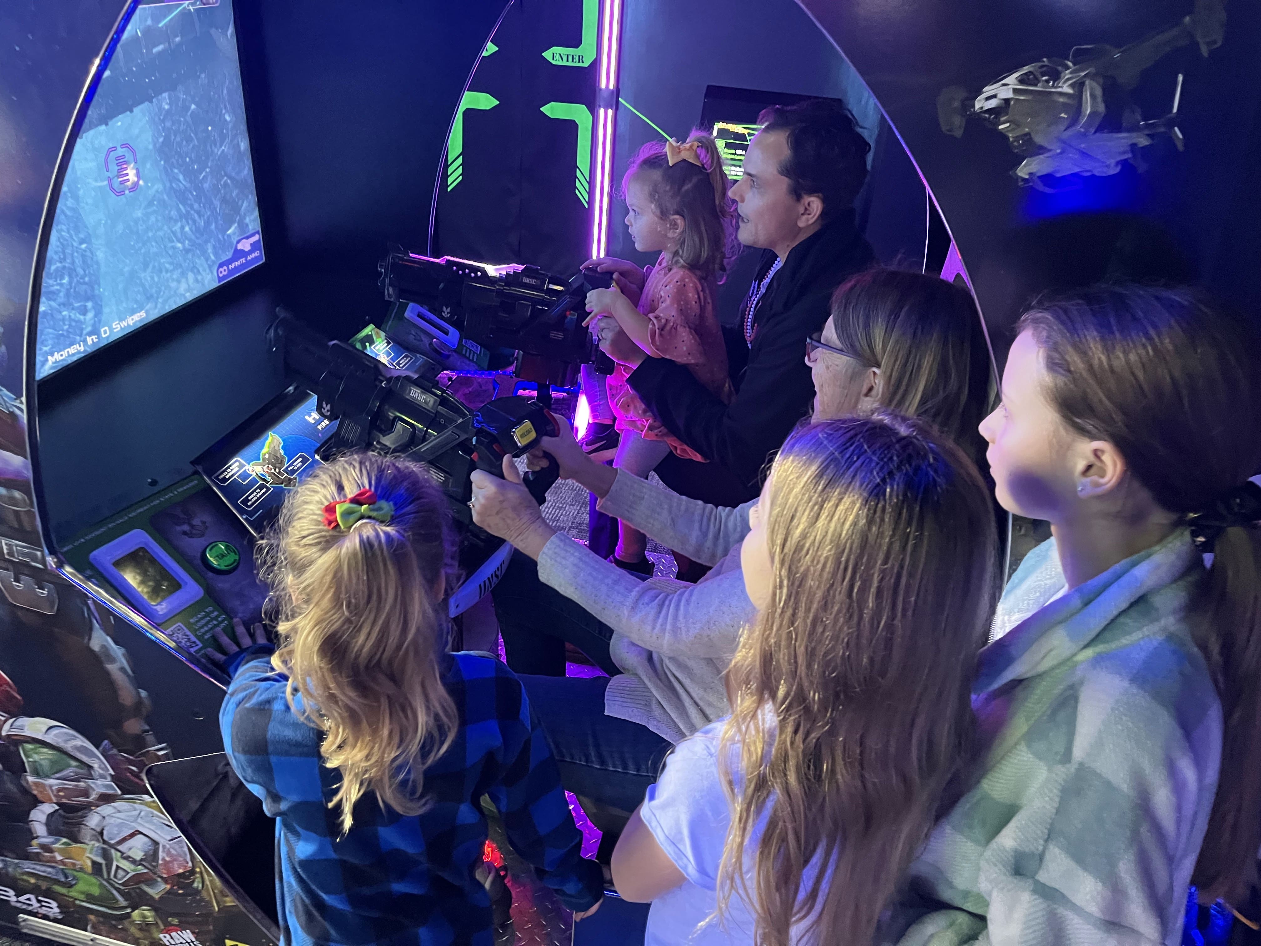 A group of children and two adults are playing an arcade shooting game together, focusing on the screen and controls in a brightly lit, futuristic game booth near Cummin's Factoria chiropractic care office location.