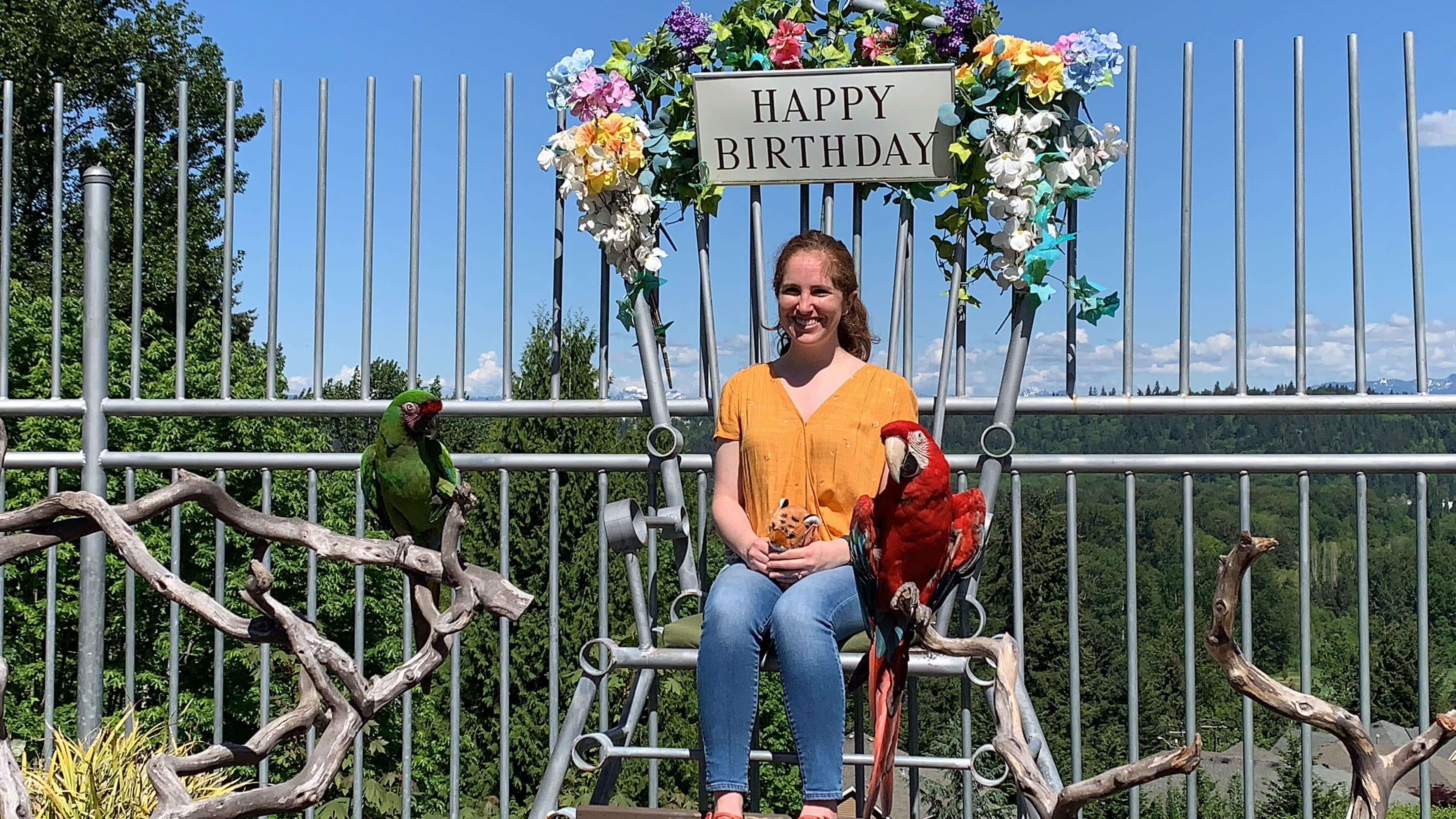 A woman in a yellow shirt sits on a large chair decorated with flowers and a Happy Birthday sign in Issaquah. Two colorful parrots perch nearby, framed by a scenic, tree-filled landscape in the background.