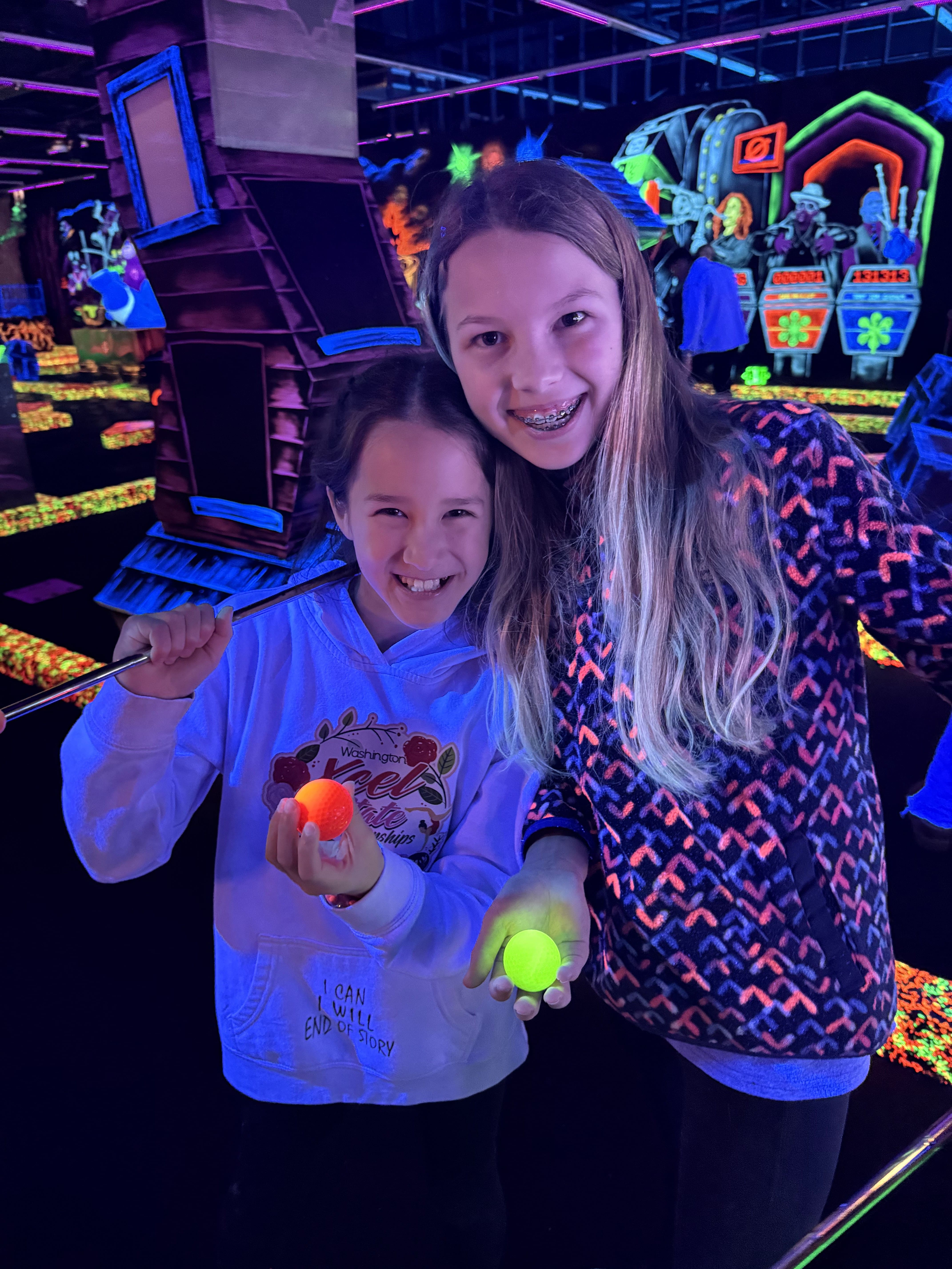 Two smiling girls stand in a glowing mini-golf course, holding neon golf balls and clubs. The vibrant, blacklight-lit decorations create a lively atmosphere—perfect for a fun day out after your visit to Cummins Factoria Chiropractic Office.