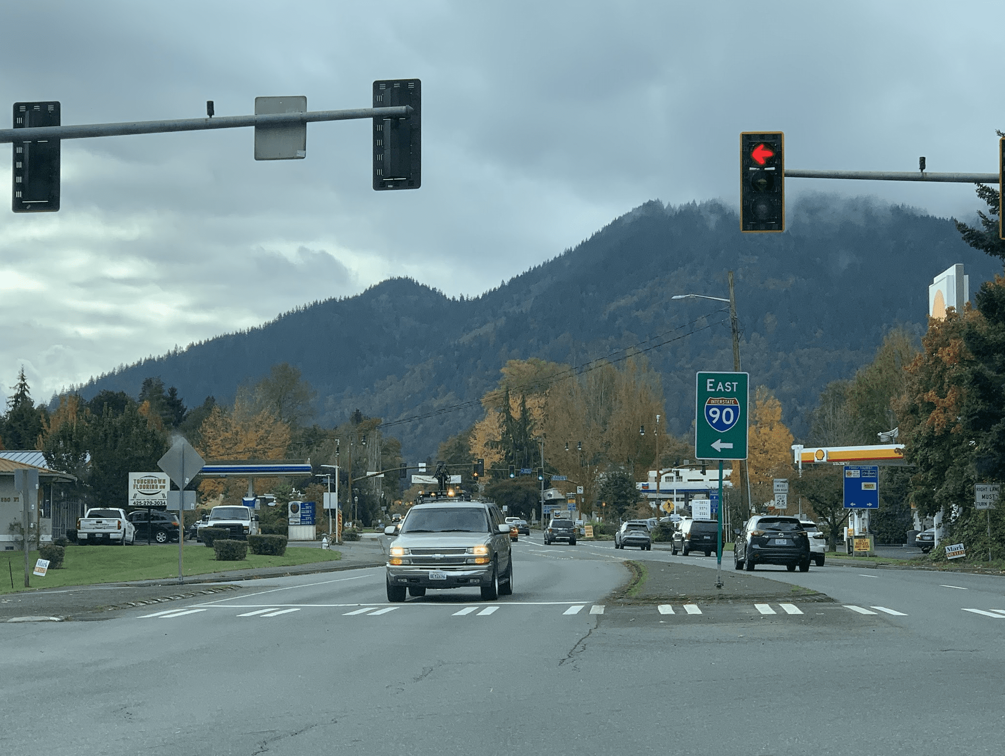An intersection in Issaquah with a red traffic light, cars waiting and driving, a green sign for I-90 East, and tree-covered mountains in the background under a cloudy sky.