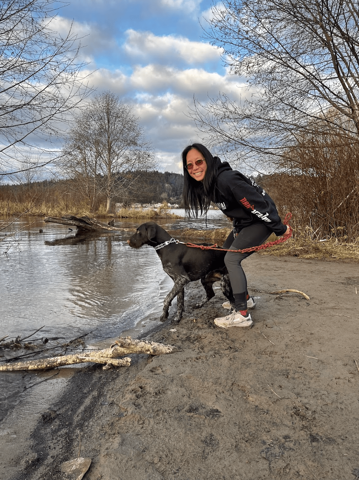 A woman in sunglasses smiles while holding a leashed dog by an Issaquah lakeshore, surrounded by bare trees and a cloudy blue sky. The dog stands near the water’s edge as the woman, in dark clothes and pink shoes, enjoys the serene setting.
