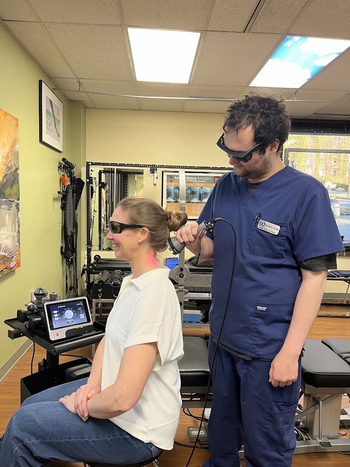 A woman sits smiling while a man in scrubs uses a laser therapy device to treat her nerd neck. Both wear protective glasses in a medical clinic with various equipment and artwork visible in the background.