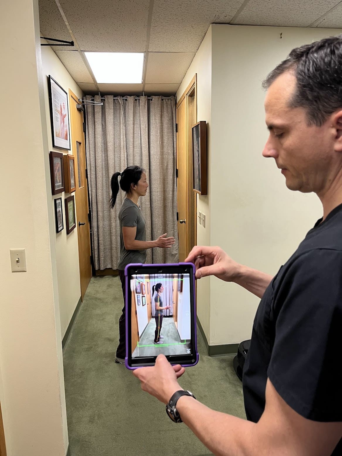 A man with slight nerd neck holds a tablet displaying a live camera view of a woman in a hallway. She appears both on the tablet screen and in the background, standing near framed pictures on the wall.