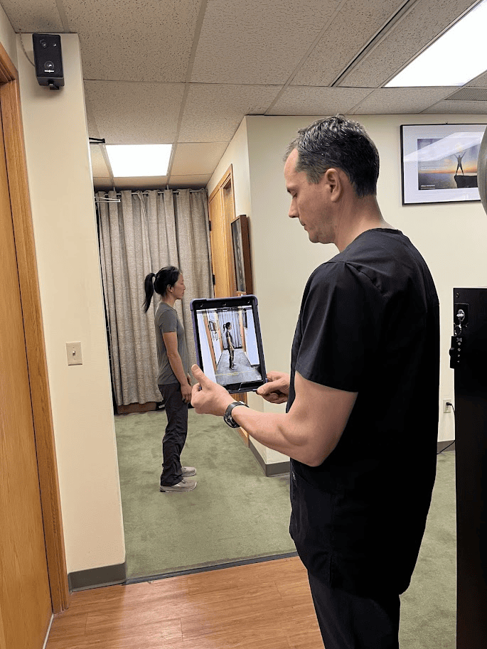 A chiropractor for migraines in scrubs takes a photo of a woman standing sideways using a tablet in a medical office with a green carpet and beige curtains.