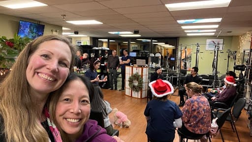 Two women smile for a selfie in the foreground at a festive indoor gathering. People in the background wear Santa hats and chat in groups. The room is decorated with holiday wreaths and string lights.