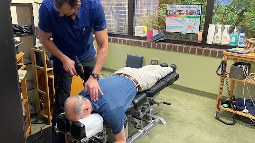 A chiropractor uses an adjustment tool on a man lying face down on a treatment table in an office with medical supplies and a large window.