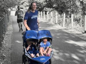 A smiling woman jogs outdoors on a paved trail while pushing a double stroller with two young children wearing blue shirts. The woman wears a “Mercer Island Bluebirds” shirt. Trees and a wooden fence line the path, and a jogger runs behind them in the background.