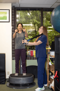 A woman stands on a Power Plate vibration platform holding resistance bands while a trainer in blue scrubs assists and smiles. They are in a fitness or physical therapy room with a large window, exercise equipment, and greenery visible outside.